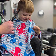 Eli is registered to the contest to win money with this photo: child, boy, haircut, barbershop, cape, octopus, comb, scissors, smile, happy, hair, stylist, chair, indoor, short_hair, person, kid, hairdresser, haircutting, fun