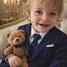 toddler, child, smiling, blue_suit, tie, teddy_bear, plush_toy, formal_wear, portrait, indoor, luxury, furniture, couch, happy, cute, blonde_hair, baby, person, seated, photography
