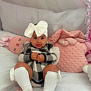 Aitana is registered to the contest to win money with this photo: baby, infant, child, headband, bow, clothing, pillow, pink, crib, soft, cute, smile, sitting, indoors, cozy, blanket, white, checkered, legs, sock