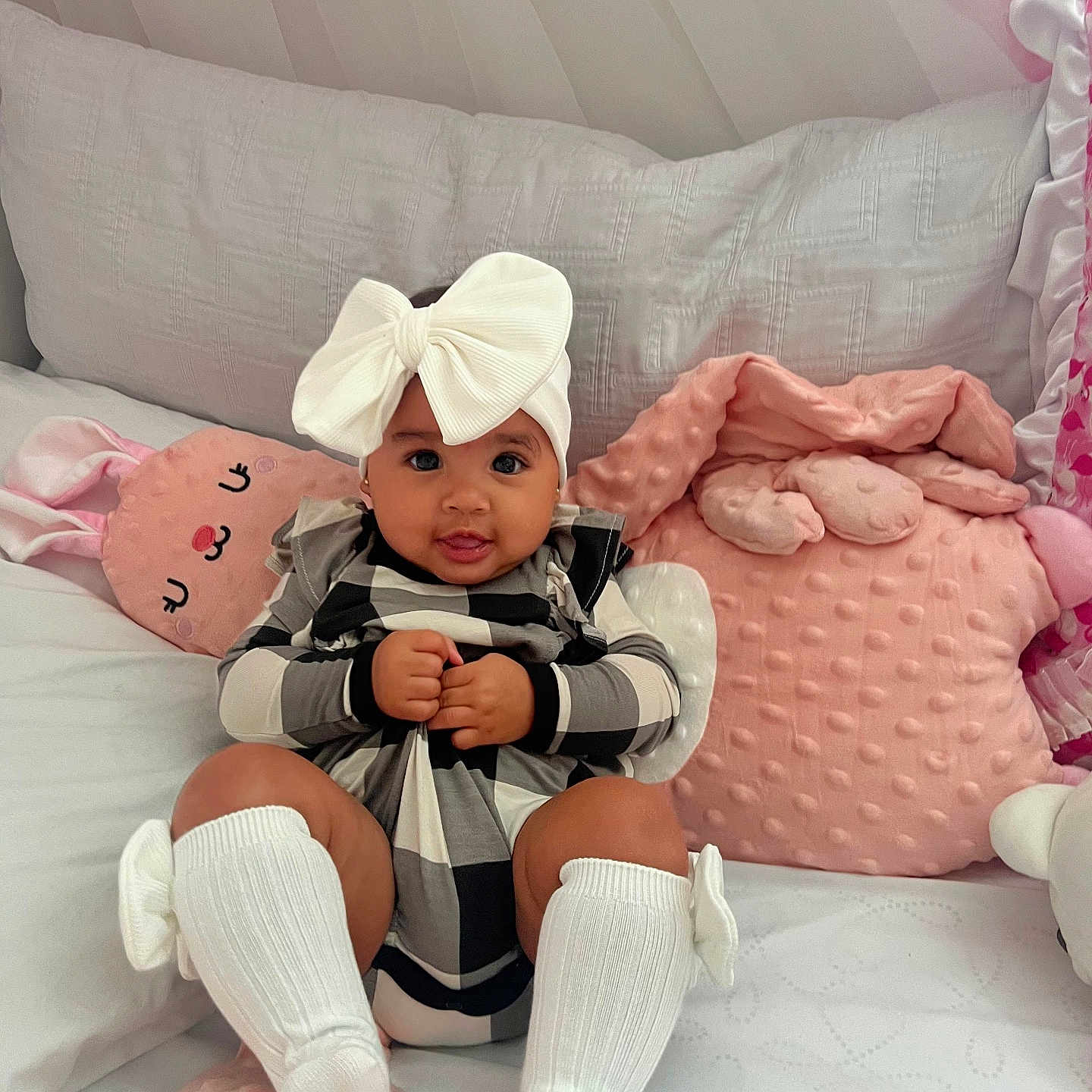 Aitana is registered to the contest to win money with this photo: baby, infant, child, headband, bow, clothing, pillow, pink, crib, soft, cute, smile, sitting, indoors, cozy, blanket, white, checkered, legs, sock