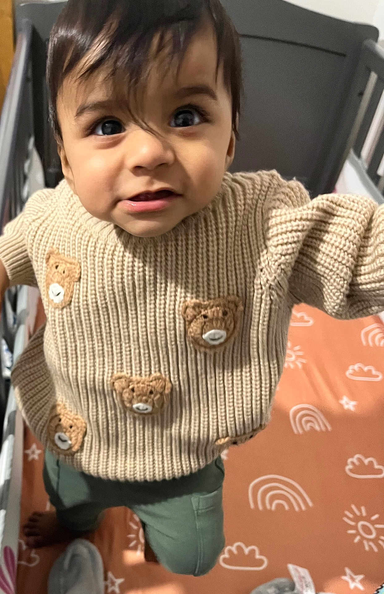Zain is registered to the contest to win money with this photo: toddler, child, sweater, knitted, teddy_bear, crib, mattress, rainbow_pattern, sun_pattern, curious, indoor, person, clothing, baby, cute, standing, close_up, face, hair, foot