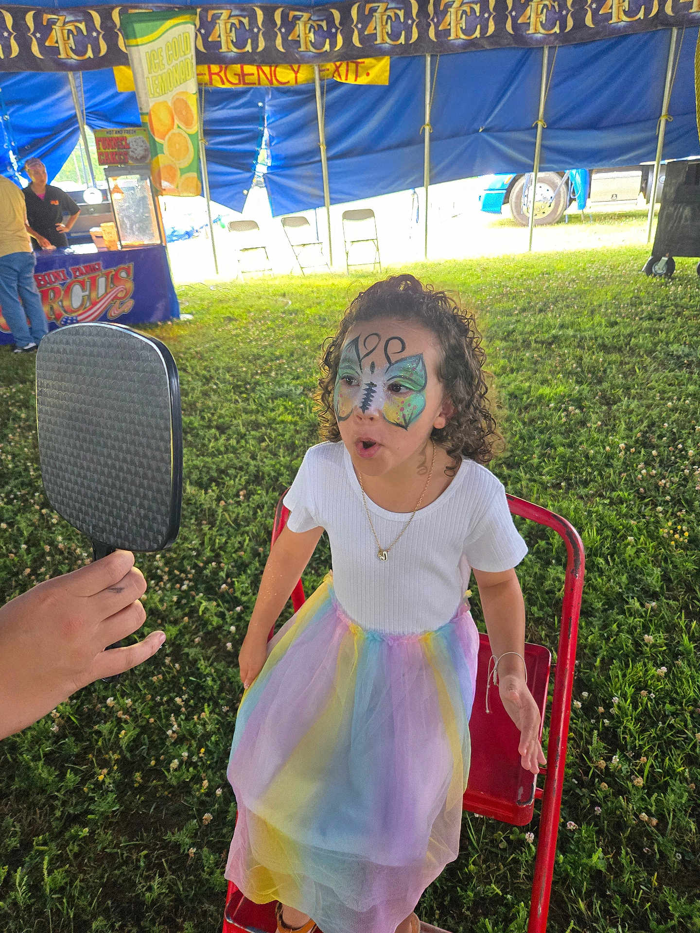 Jemma is registered to the contest to win money with this photo: child, face_paint, butterfly, curly_hair, white_shirt, rainbow_skirt, red_chair, mirror, grass, tent, circus, sign, emergency_exit, outdoor, festival, people, hand, expression, summer, event