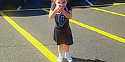 Jemma is registered to the contest to win money with this photo: child, girl, parking_lot, car, vehicle, curly_hair, white_bow, black_dress, sneakers, sunny, outdoor, asphalt, yellow_lines, standing, person, woman, casual, daytime, shadow, holding_object