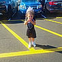 child, girl, parking_lot, car, vehicle, curly_hair, white_bow, black_dress, sneakers, sunny, outdoor, asphalt, yellow_lines, standing, person, woman, casual, daytime, shadow, holding_object