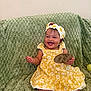 Daniella is registered to the contest to win money with this photo: baby, bright, cheerful, child, clothing, cute, daisy, footwear, green_blanket, happy, headband, indoor, infant, person, portrait, sitting, smiling, texture, wooden_sign, yellow_dress