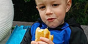Kyron participe au concours pour gagner de l'argent avec cette photo : child, boy, sandwich, food, outdoor, bench, greenery, leaves, blue_bag, jacket, black_clothing, hands, face, portrait, casual, daylight, nature, sitting, cute, young