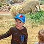 child, boy, smiling, cap, spiderman, lion, zoo, glass, enclosure, rocks, grass, toy, girl, standing, outdoor, nature, animal, fence, happy, casual_clothing