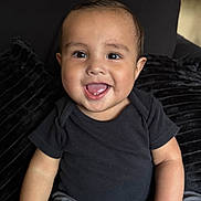Kai joined the competition — help win amazing prizes! baby, child, smiling, happy, face, sitting, couch, black_shirt, gray_pants, plush_cushion, indoors, portrait, young_child, cute, innocence, joy, person, head, eyes, hand