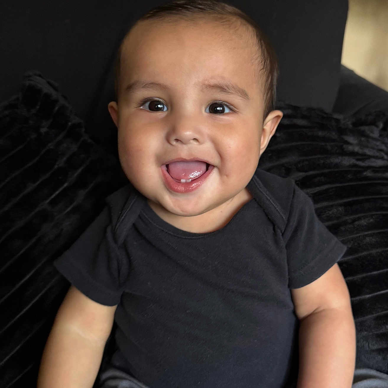 Kai joined the competition — help win amazing prizes! baby, child, smiling, happy, face, sitting, couch, black_shirt, gray_pants, plush_cushion, indoors, portrait, young_child, cute, innocence, joy, person, head, eyes, hand