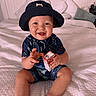 baby, child, smiling, hat, bucket_hat, blue_clothing, shark_pattern, sitting, bed, white_bedspread, pillow, indoors, cute, happy, infant, person, holding, carton, playful, portrait