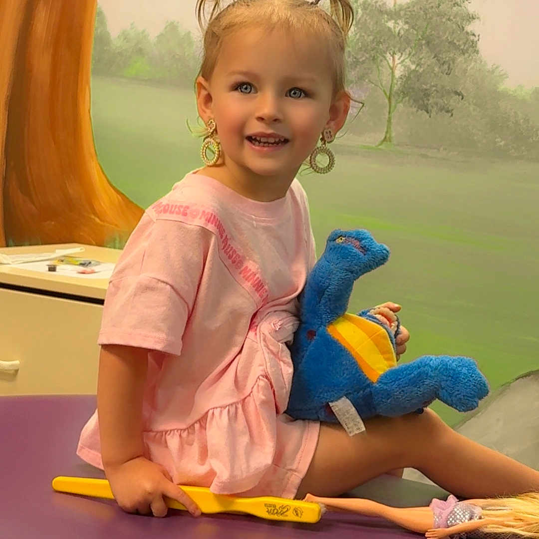 Brynnslee is registered to the contest to win money with this photo: child, girl, pink_tshirt, pigtails, earrings, plush_toy, blue_dinosaur, doll, bench, purple_surface, smiling, indoor, painting_background, tree, greenery, playful, happy, portrait, cute, toddler