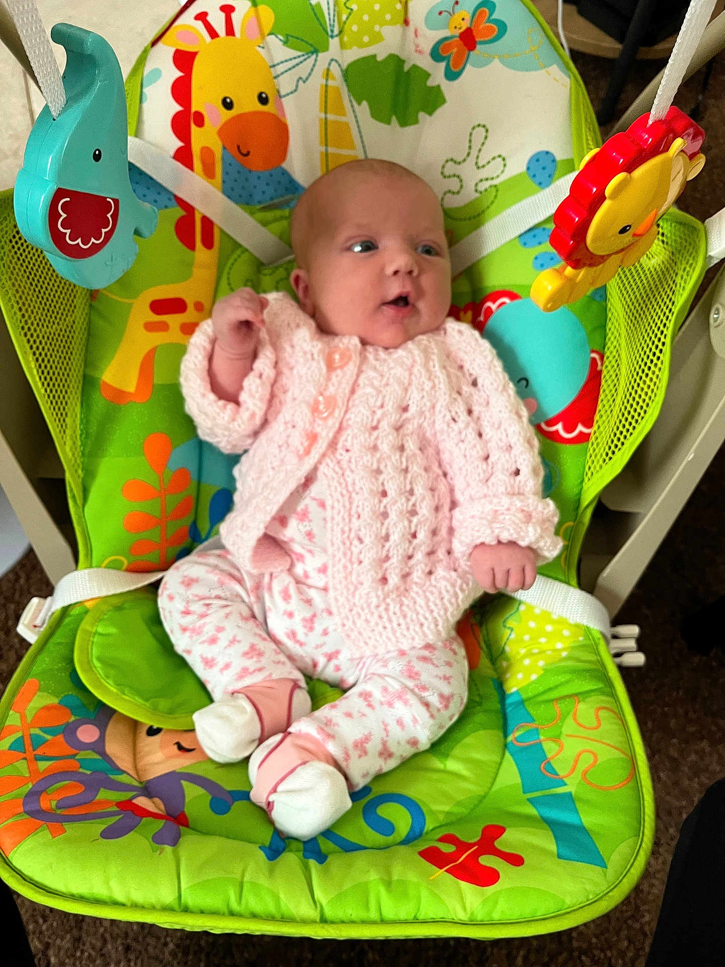 Freya is registered to the contest to win money with this photo: animal_print, baby, baby_rocker, carpet, child, colorful, comfortable, curious, cute, doll, floral_pants, indoor, infant, knitted_clothing, looking_away, pink_sweater, seated, small_hands, toys, white_socks