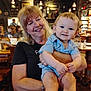 Maverick is registered to the contest to win money with this photo: baby, child, woman, smile, happy, restaurant, indoor, people, holding, casual_clothing, blonde_hair, baby_boy, seated, portrait, warm_lighting, background, blurred, furniture, table, chairs