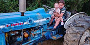 Maverick is registered to the contest to win money with this photo: blue, casual_clothing, child, dirt, footwear, ford, fun, grass, greenery, happy, hugging, nature, outdoor, playful, rustic, siblings, smiling, summer, tractor, vehicle