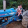 child, tractor, ford, blue, outdoor, grass, greenery, smiling, siblings, hugging, vehicle, rustic, playful, fun, nature, summer, casual_clothing, footwear, dirt, happy