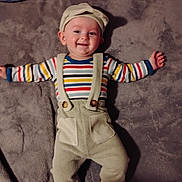 Maverick is registered to the contest to win money with this photo: baby, child, smile, striped_shirt, overalls, hat, blanket, soft_texture, lying_down, happy, infant, indoors, cute, face, headwear, clothing, person, baby_clothes, socks, portrait