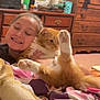 Kilo is registered to the contest to win money with this photo: person, cat, indoor, smiling, hugging, furniture, dresser, blanket, orange_cat, white_cat, bedroom, clutter, happy, legs_up, cozy, casual, relaxed, pet, feline, human