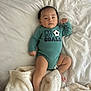 baby, infant, onesie, text_on_clothing, soccer_ball_graphic, blanket, bed, white_bed_sheet, soft_texture, lying_down, child, skin, face, legs, arms, foot, hand, casual_clothing, indoor, calm_expression