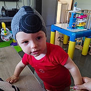 Esteban participe au concours pour gagner de l'argent avec cette photo : adult_hand, beanie, child, colorful, curious, face, floor, furniture, hand, hat, indoor, leaning, person, plastic_toy, playroom, red_clothing, table, toddler, toy, young_child