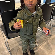 Brandon joined the competition — help win amazing prizes! child, indoor, dreadlocks, green_jacket, ripped_clothing, pants, black_shoes, toy, candy, holding, standing, curious, person, floor, chair, black_chair, casual_clothing, young, expression, face