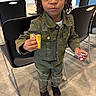 child, indoor, dreadlocks, green_jacket, ripped_clothing, pants, black_shoes, toy, candy, holding, standing, curious, person, floor, chair, black_chair, casual_clothing, young, expression, face