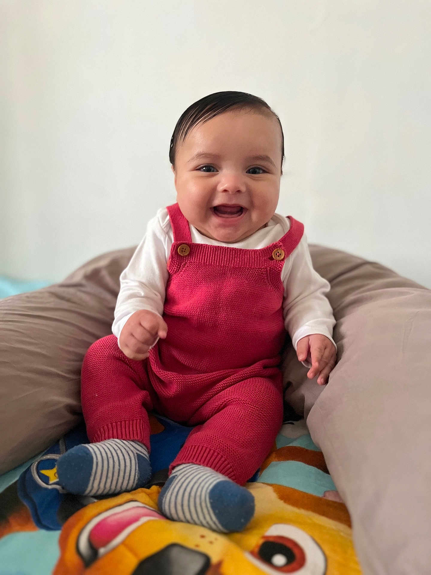 Timéo a rejoint le concours — aidez-le/la à gagner de superbes lots ! baby, smiling, red_overalls, white_shirt, cushion, blanket, striped_socks, indoor, child, happy, cute, portrait, seated, infant, knitwear, colorful, soft_furniture, playful, closeup, young_child