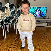 Timothée participe au concours pour gagner de l'argent avec cette photo : child, boy, smile, standing, sneakers, white_pants, sweatshirt, indoor, wooden_floor, television, furniture, chair, leaf_pattern, hands_in_pockets, person, home, casual_clothing, short_hair, happy, living_room