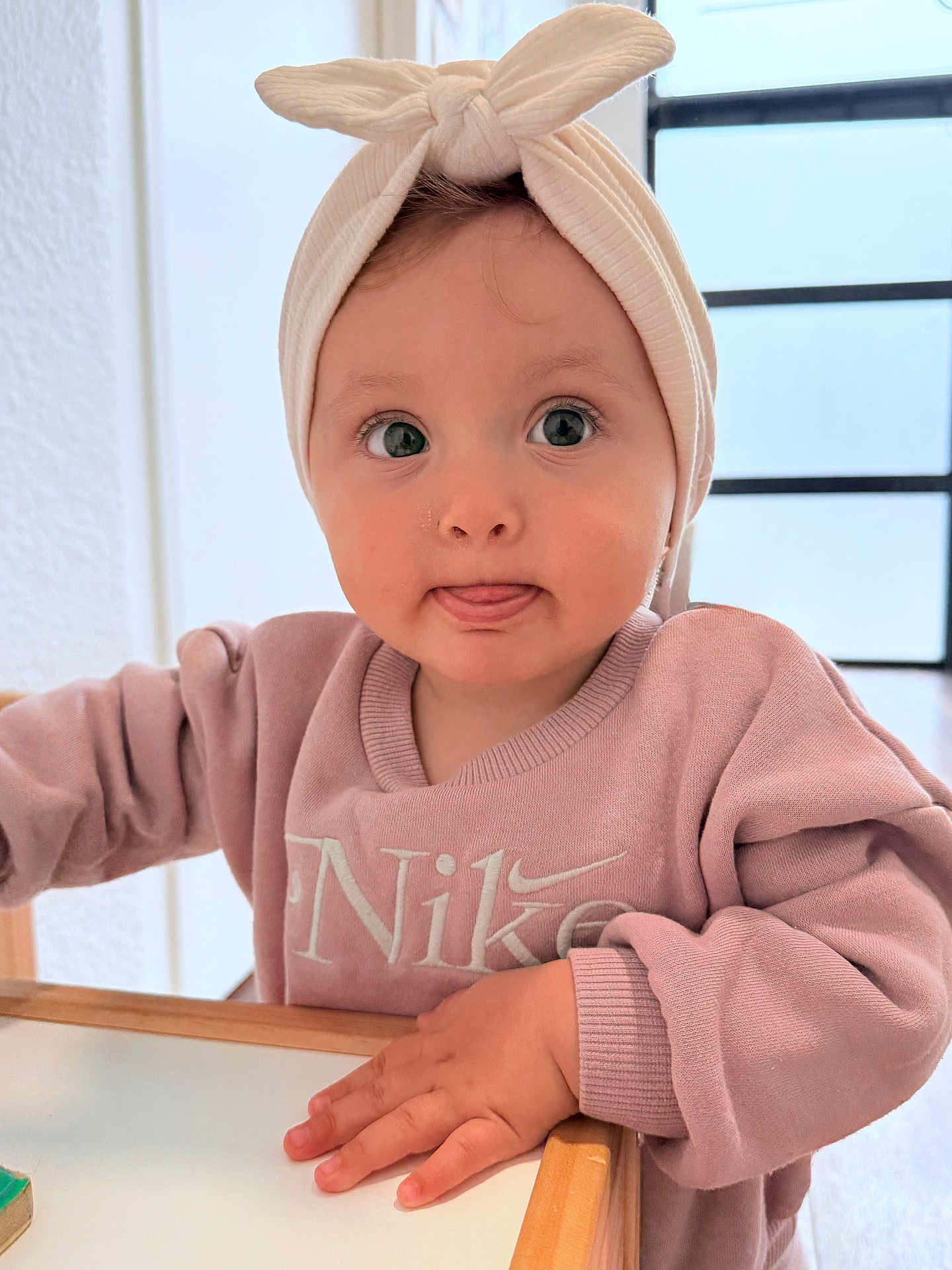 Victoire participe au concours pour gagner de l'argent avec cette photo : baby, child, headband, pink_sweatshirt, nike, tongue_out, hand, table, wooden_table, indoor, curious, face, large_eyes, sitting, sweater, long_sleeves, natural_light, person, closeup, cute