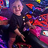 toddler, child, arcade, arcade_machine, motorcycle_ride, racing_game, neon_lights, colorful, smiling, portrait, indoor, entertainment_center, game_room, seated, black_shirt, sandals, toy_vehicle, family_outing, bright_lighting, recreation