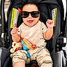 baby, child, car_seat, sunglasses, smiling, toy, bib, clothing, infant, happy, cute, hands, seatbelt, plaything, casual, indoors, portrait, young_child, accessory, comfort