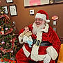 santa_claus, baby, christmas_tree, holiday, festive, red_suit, white_beard, glasses, decorations, candy_cane, gift_box, indoor, smiling, child, person, celebration, warm_lighting, wooden_chair, wall_art, wifi_sign