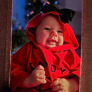 Olivia is registered to the contest to win money with this photo: baby, black_bow, blurred_background, child, christmas_lights, cozy, cute, face, festive, framed, hands, holiday, hood, indoors, infant, portrait, red_clothing, smiling, soft_lighting, warm_colors