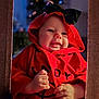 baby, black_bow, blurred_background, child, christmas_lights, cozy, cute, face, festive, framed, hands, holiday, hood, indoors, infant, portrait, red_clothing, smiling, soft_lighting, warm_colors