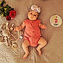 baby, infant, flower_headband, coral_romper, plush_toy, elephant_toy, wooden_teether, red_roses, quilted_bedspread, birthday_badge, milestone, lying_down, cute, child, portrait, baby_boy, baby_girl, soft_lighting, indoor, celebration