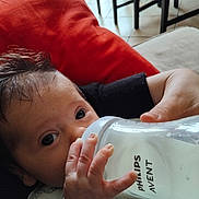 Mathéo a rejoint le concours — aidez-le/la à gagner de superbes lots ! baby, infant, milk_bottle, feeding, hand, face, eyes, couch, cushion, red_cushion, indoors, person, clothing, patterned_outfit, milk, close_up, floor, furniture, adult_hand, bottle