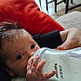 baby, infant, milk_bottle, feeding, hand, face, eyes, couch, cushion, red_cushion, indoors, person, clothing, patterned_outfit, milk, close_up, floor, furniture, adult_hand, bottle