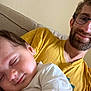 baby, child, man, glasses, sleeping, smile, yellow_shirt, white_clothing, couch, indoor, person, face, beard, closeup, portrait, rest, comfort, family, love, casual
