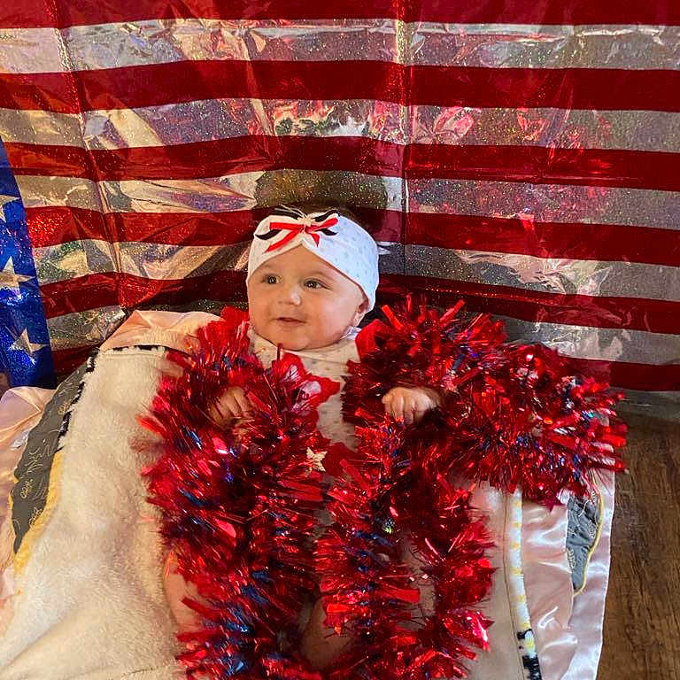 Chevelle joined the competition — help win amazing prizes! american_flag, baby, blanket, blue, child, cute, decorations, festive, floor, headband, holiday, indoors, patriotic, red, sitting, smiling, soft, tinsel, white, wrapped