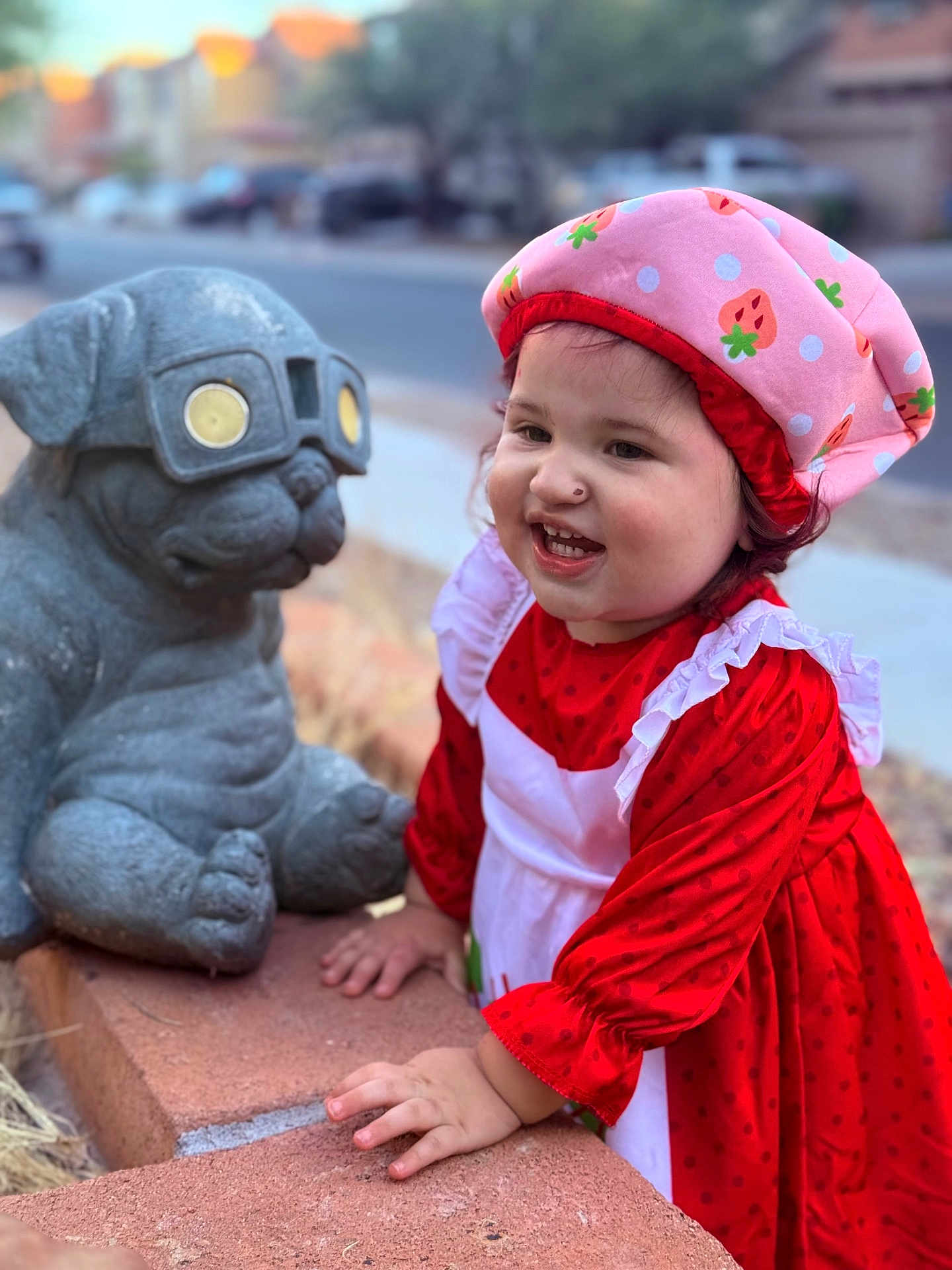 Elleanna joined the competition — help win amazing prizes! toddler, child, red_dress, pink_hat, strawberry_pattern, smiling, happy, stone_statue, pug_statue, glasses, brick_ledge, outdoor, suburban, daylight, portrait, cute, playful, person, side_view, young_child