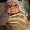baby, infant, hat, onesie, floral_pattern, wide_eyes, face, lying_down, blanket, quilt, comfort, cute, indoors, child, newborn, soft_lighting, portrait, expression, bed, clothing