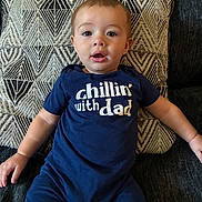 Roman is registered to the contest to win money with this photo: baby, blanket, blue_outfit, centered_composition, child, close_up, couch, cushion, family_photo, furniture, geometric_pattern, indoors, infant, open_mouth, portrait, relaxed, sitting, t_shirt, text_on_clothing, wide_eyed