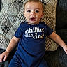 baby, blanket, blue_outfit, centered_composition, child, close_up, couch, cushion, family_photo, furniture, geometric_pattern, indoors, infant, open_mouth, portrait, relaxed, sitting, t_shirt, text_on_clothing, wide_eyed