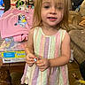 toddler, child, indoor, dress, pastel_colors, chalk, toy, pink_sweatshirt, cartoon_character, furniture, couch, wooden_table, messy_hair, person, floor, clutter, hand, face, smile, young_child