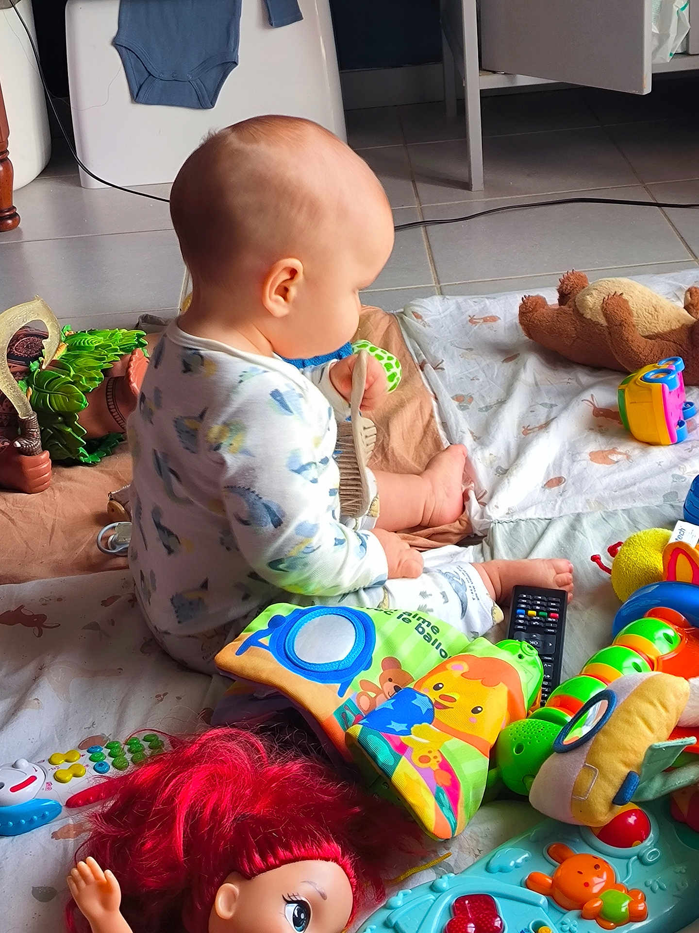Ewen participe au concours pour gagner de l'argent avec cette photo : baby, child, toy, doll, brush, floor, blanket, colorful, indoor, play, plaything, toddler, red_hair, remote_control, stuffed_animal, patterned_blanket, clothing, sitting, curious, head