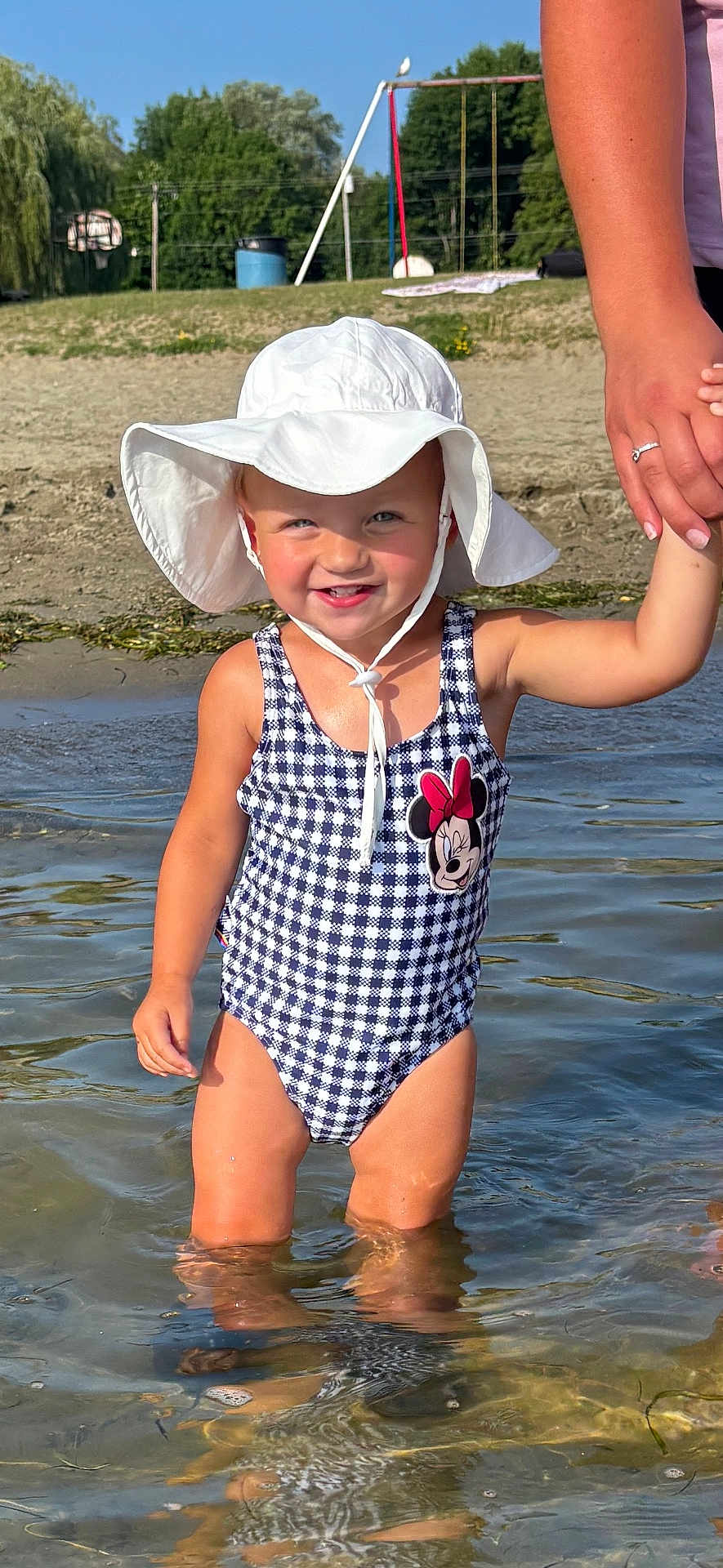 Emma is registered to the contest to win money with this photo: toddler, child, water, swimsuit, hat, sun_hat, holding_hand, beach, outdoor, summer, playground, sand, smiling, person, sunny, nature, trees, water_reflection, minnie_mouse, happy