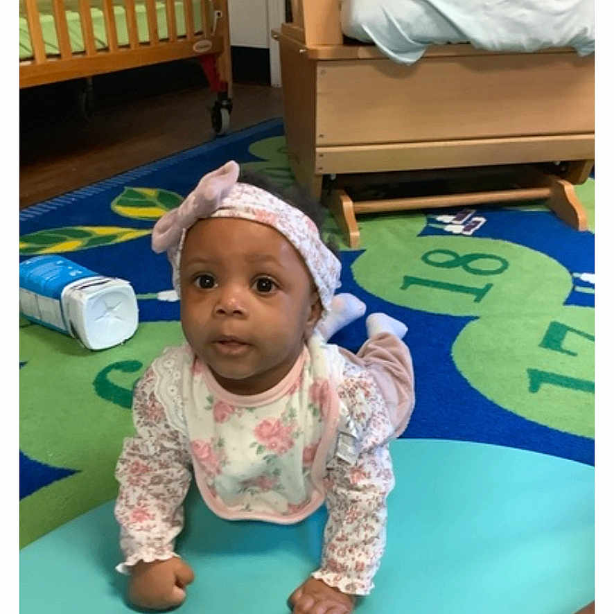 Alani is registered to the contest to win money with this photo: baby, infant, child, headband, bib, floor_mat, playroom, carpet, wooden_furniture, curious, hands, crawling, indoor, colorful, young_child, person, cute, childrens_room, soft_surface, alert