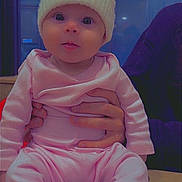 Skyla-Rae is registered to the contest to win money with this photo: baby, child, infant, hat, knit_hat, pink_clothing, socks, hands, wooden_table, indoor, person, cute, seated, face, eyes, apparel, warm_clothing, holding, background_sign, portrait