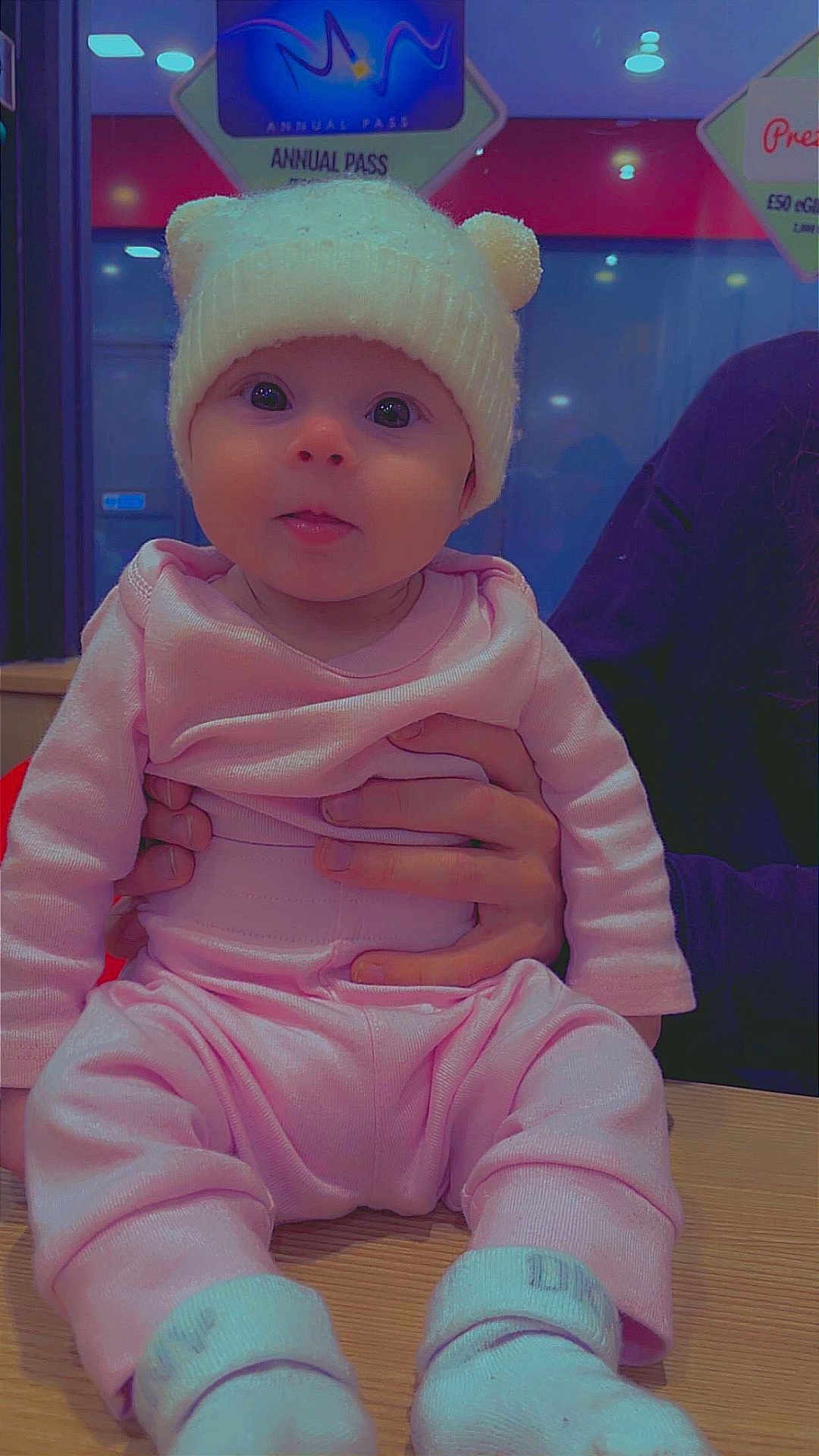 Skyla-Rae is registered to the contest to win money with this photo: baby, child, infant, hat, knit_hat, pink_clothing, socks, hands, wooden_table, indoor, person, cute, seated, face, eyes, apparel, warm_clothing, holding, background_sign, portrait