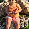 toddler, child, outdoor, rock, sunlight, greenery, flowers, orange_tank_top, striped_shorts, sandals, curious_expression, moss, nature, summer, portrait, sitting, baby, cute, daylight, person