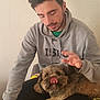 man, dog, indoor, pet, hoodie, brown_dog, sitting, lap, tongue_out, casual, relaxed, home, facial_hair, short_hair, textured_wall, black_pants, blanket, affection, friendship, comfort